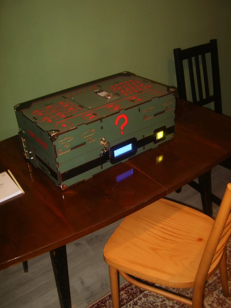 Zielona, elektroniczna skrzynia z czerwonym znakiem zapytania i podświetlanymi przyciskami na stole w escape roomie.