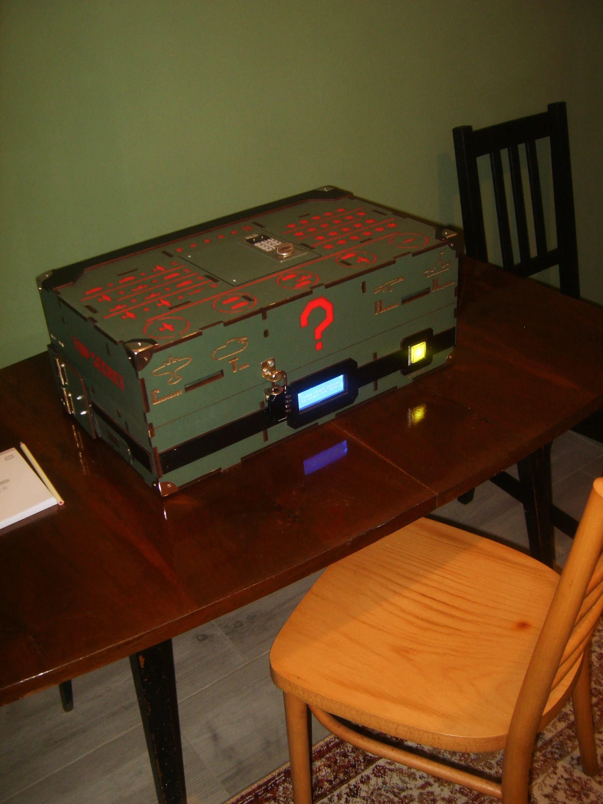 Zielona, elektroniczna skrzynia z czerwonym znakiem zapytania i podświetlanymi przyciskami na stole w escape roomie.