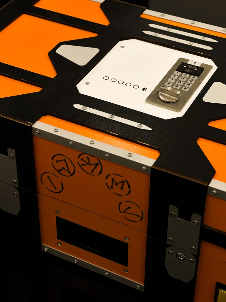 Elektroniczna skrzynia z zamkiem szyfrowym i zagadkowymi symbolami, używana jako element gry w escape roomie.