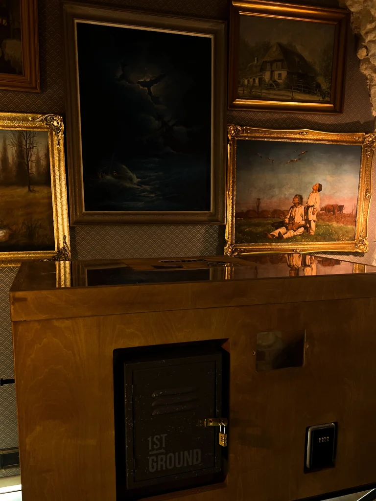 Zamknięta skrzynia z napisem "1st Ground" na tle ściany z obrazami, element gry w escape roomie o klimacie tajemncia wawelu